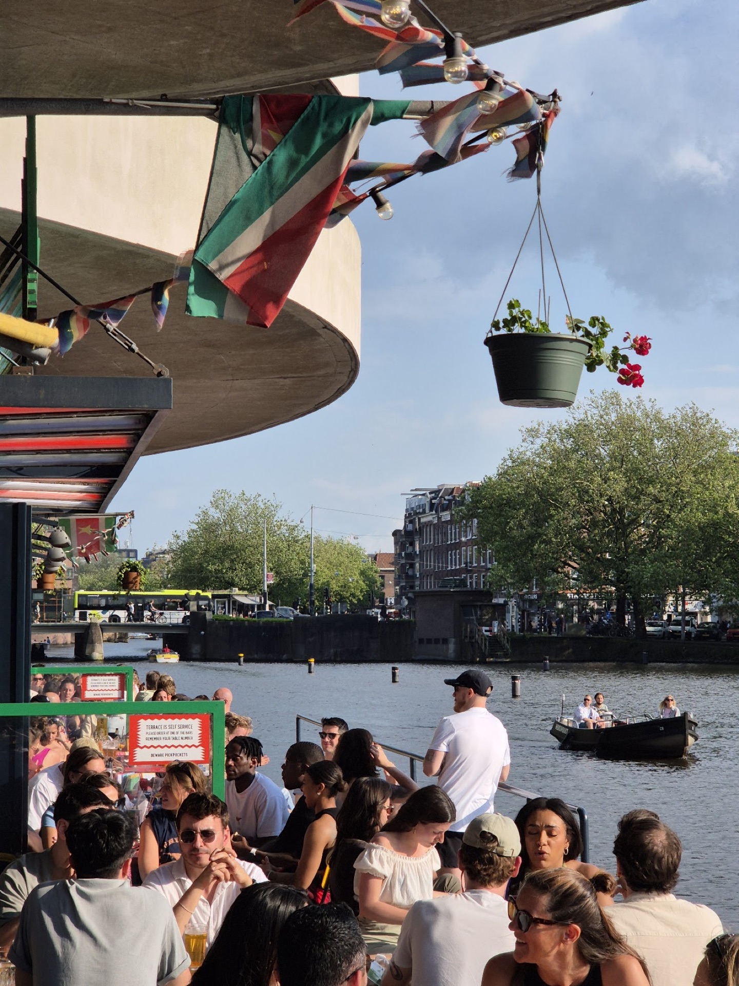 Terraço lotado do Waterkant num dia de sol, com um barquinho de aluguel passando no Amstel ao fundo